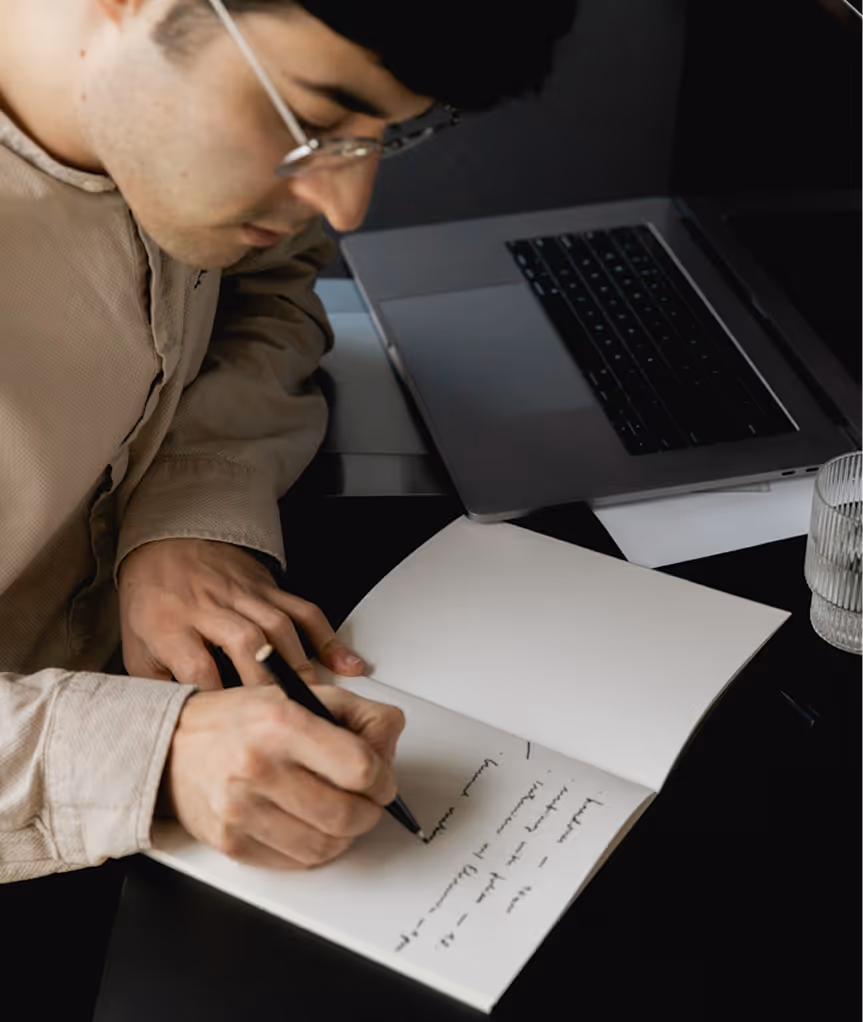 Homme écrivant dans un carnet blanc, ordinateur portable fermé et verre d'eau à côté.
