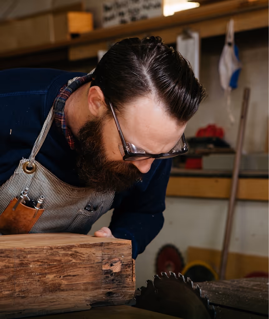 Un homme barbu portant un tablier inspecte un morceau de bois au-dessus d'une scie circulaire en atelier.
