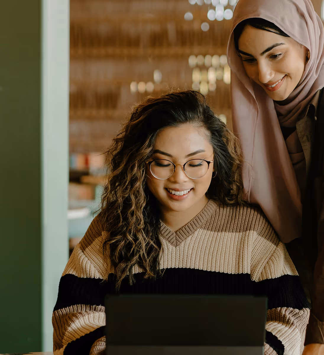 Deux femmes regardent un ordinateur portable, l'une sourit portant un hijab rose pâle.