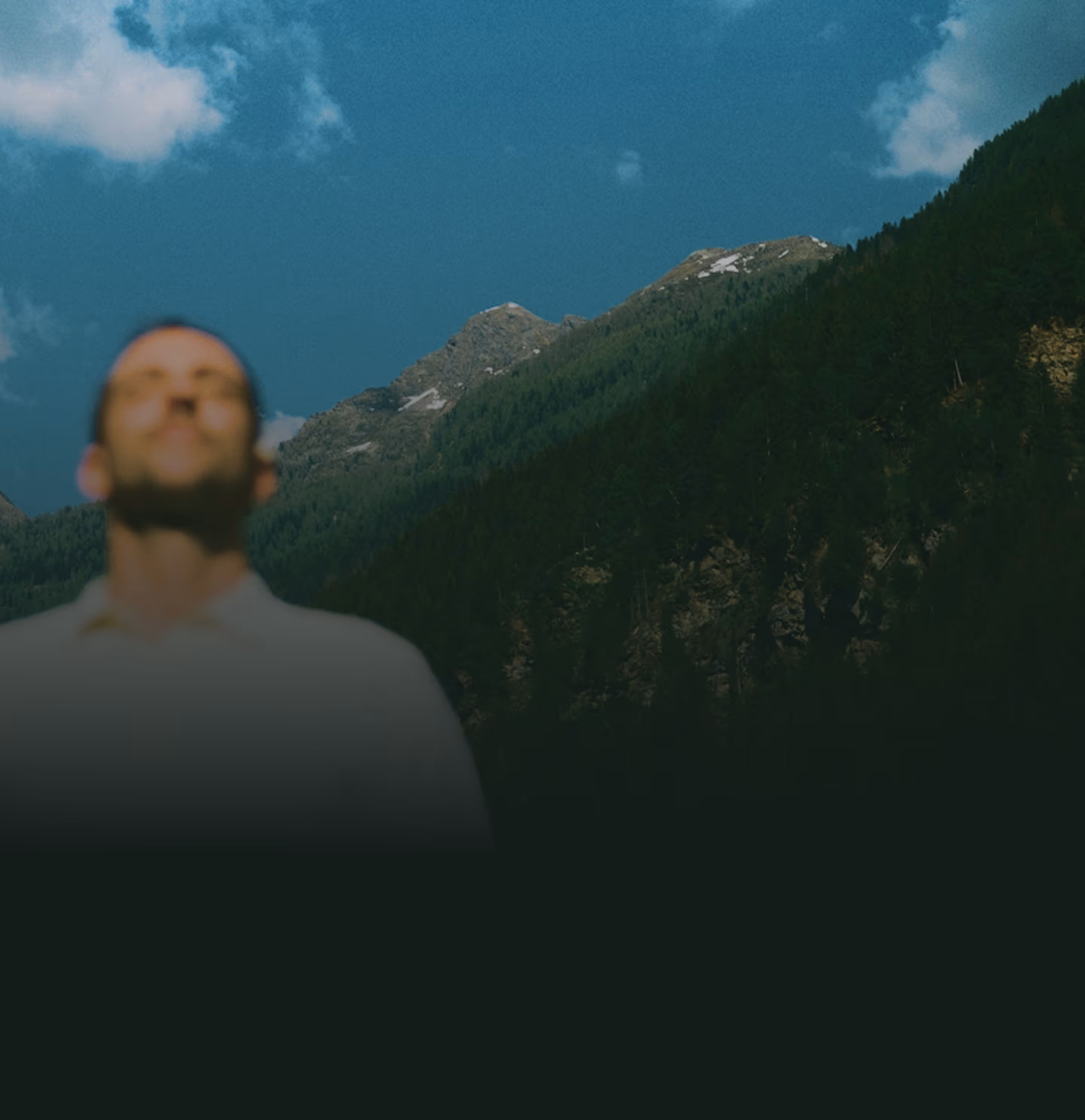 Homme flou les yeux fermés devant une montagne boisée sous un ciel bleu avec quelques nuages.