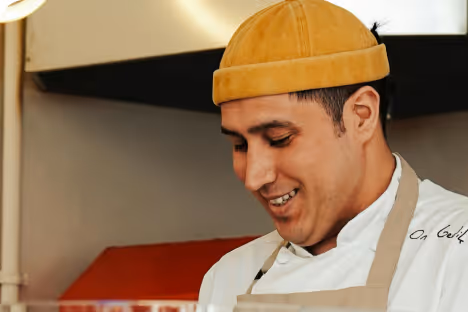 Un chef souriant portant une toque jaune et un tablier beige dans une cuisine.