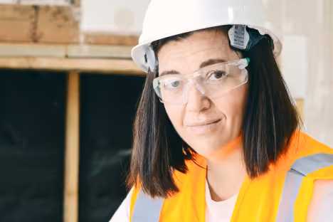Femme portant un casque blanc, des lunettes de protection et un gilet de sécurité orange sur un chantier.