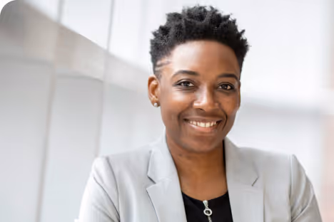 Femme souriante en veste grise posant près d'une fenêtre dans un environnement lumineux.