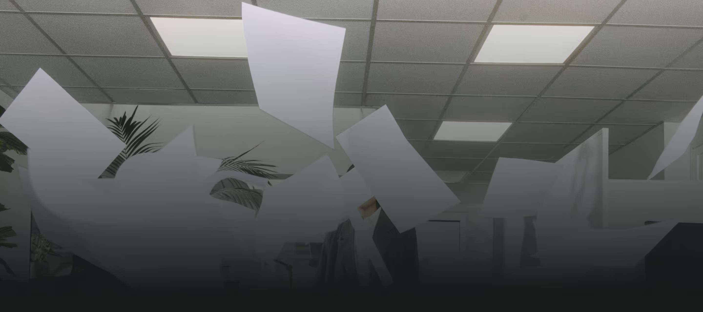 Des feuilles de papier volent dans un bureau moderne sous un plafond avec des luminaires encastrés.