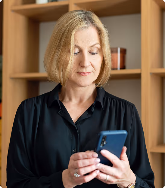 Femme blonde portant une chemise noire regardant un smartphone dans une pièce avec une étagère en bois.