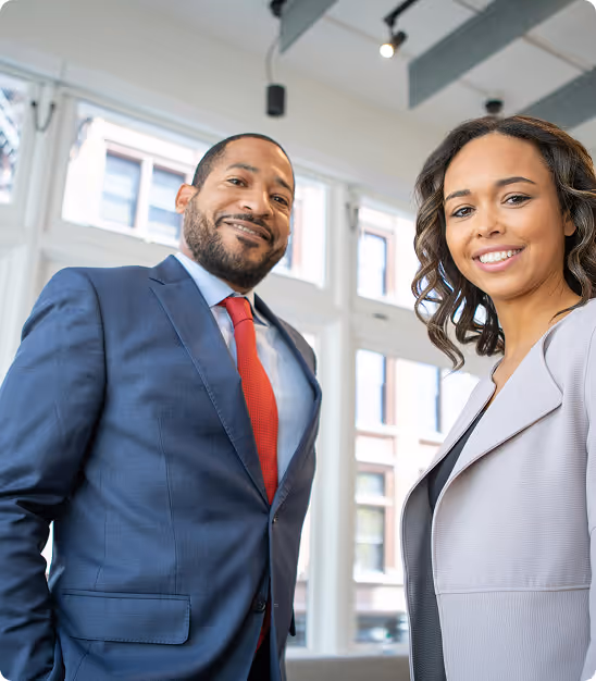 Deux professionnels souriants en tenue d'affaires dans un bureau moderne aux grandes fenêtres.