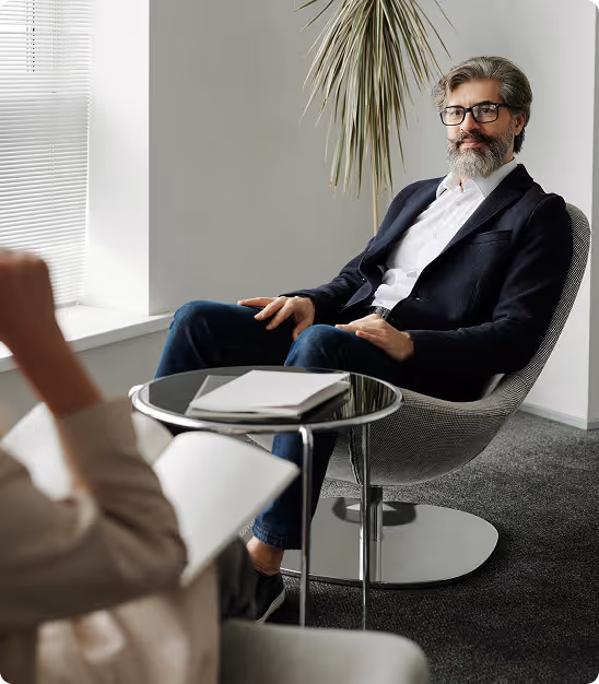 Homme avec barbe et lunettes assis dans un fauteuil avec table ronde entre deux personnes en discussion.