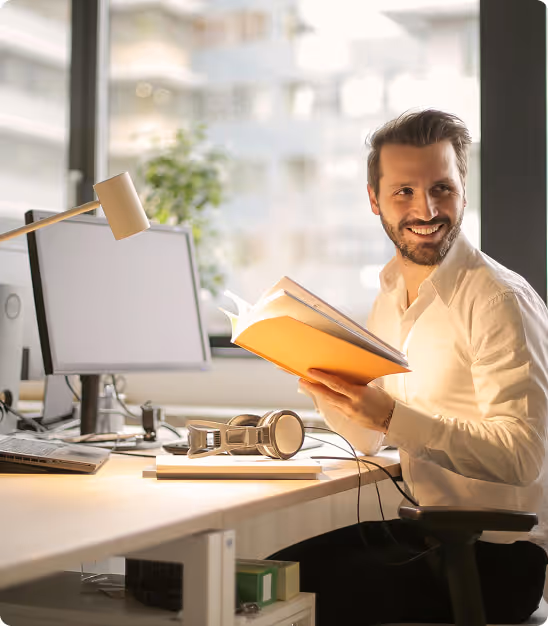 Jeune homme souriant lisant un livre à son bureau avec ordinateur, casque, et lampe.