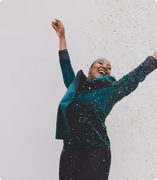 Femme souriante en veste verte lançant des confettis avec joie sur fond gris.