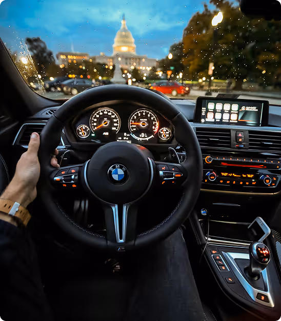 Vue du volant d'une BMW avec tableau de bord éclairé, route mouillée et bâtiment au loin au crépuscule.