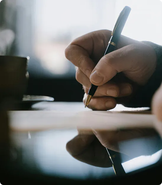 Main écrivant avec un stylo noir sur une feuille, reflet visible sur une surface brillante.
