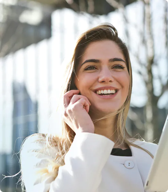 Jeune femme souriante portant un manteau blanc, tenant un appareil, en extérieur au soleil.