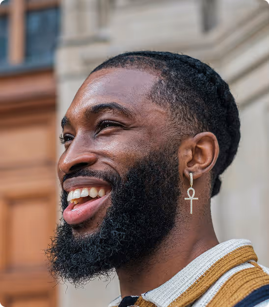 Homme souriant avec barbe et boucle d'oreille en forme d'ankh, en extérieur devant un bâtiment.
