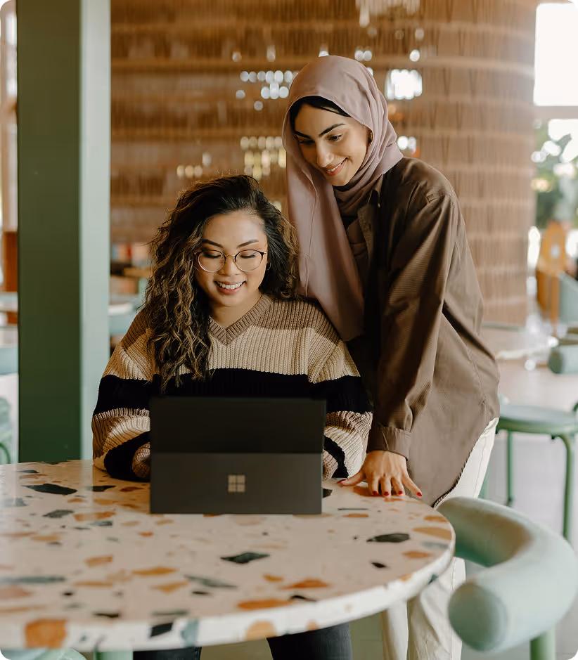 Deux femmes souriantes utilisant un ordinateur portable à une table ronde dans un café lumineux.