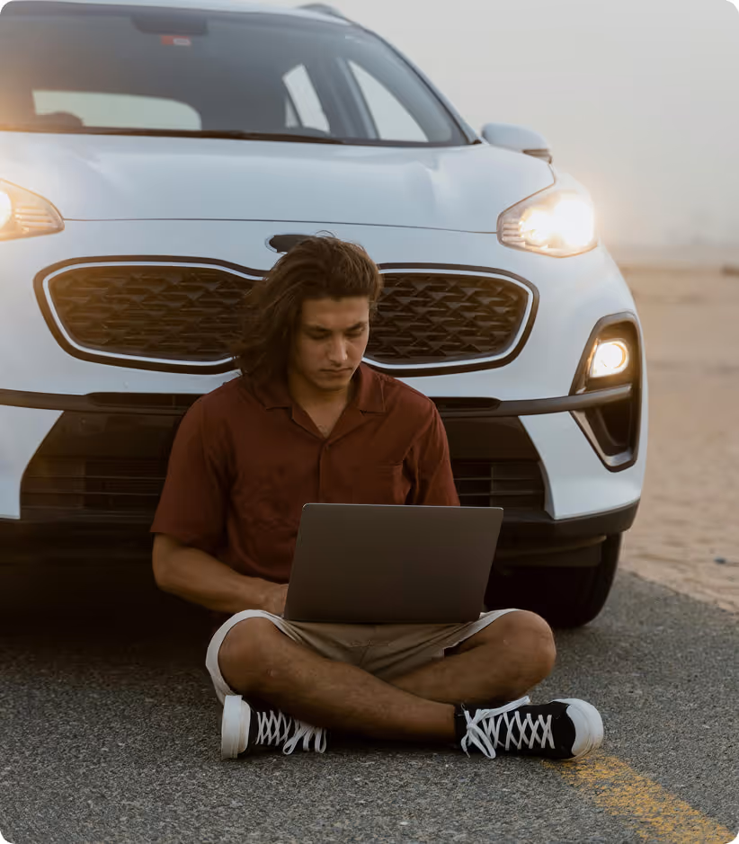 Un homme assis par terre devant une voiture blanche allumée utilisant un ordinateur portable.