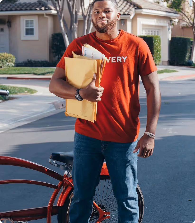 Homme souriant tenant des enveloppes debout devant un vélo rouge dans un quartier résidentiel.