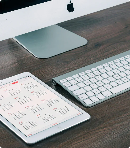 Ordinateur Apple, clavier et tablette affichant un calendrier annuel sur une surface en bois.
