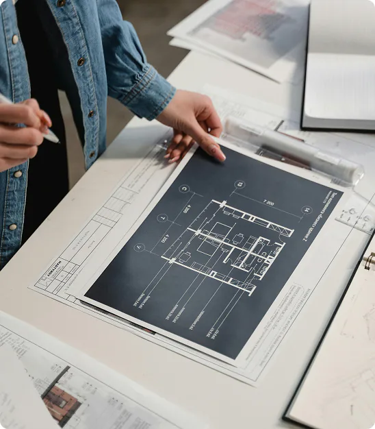Person holding pen reviewing architectural floor plan and blueprints on a white desk.