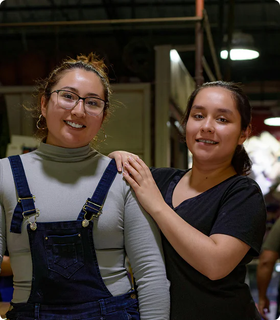 Deux femmes souriantes, l'une en salopette bleue et col roulé gris, l'autre en t-shirt noir, posant ensemble.