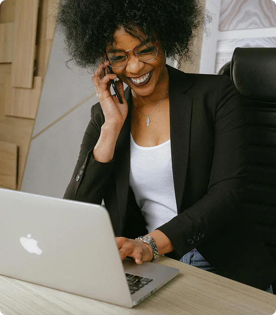 Femme souriante en lunettes parlant au téléphone tout en travaillant sur un ordinateur portable Apple.