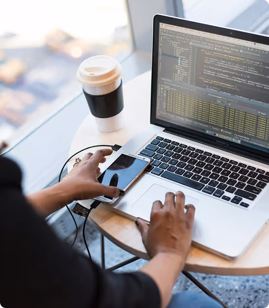 Une personne utilise un ordinateur portable affichant du code avec un téléphone et un café à côté.