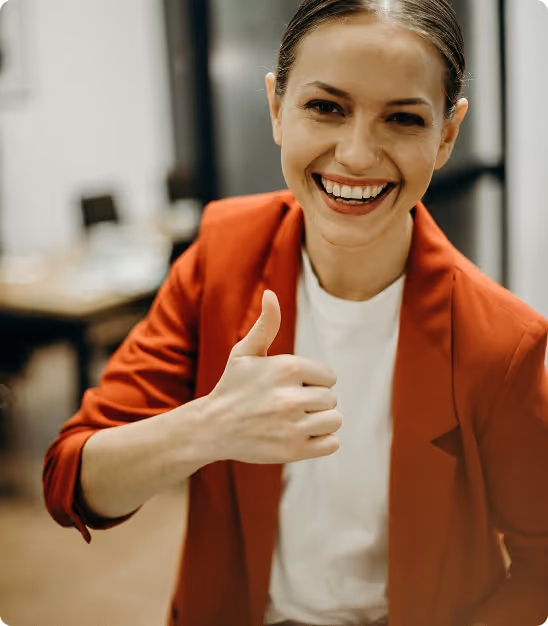 Femme souriante en veste rouge montrant un pouce levé dans un bureau flou.