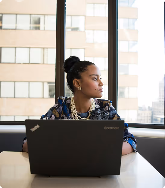 Femme d'affaires portant un collier de perles, regardant par la fenêtre derrière un ordinateur portable Lenovo.