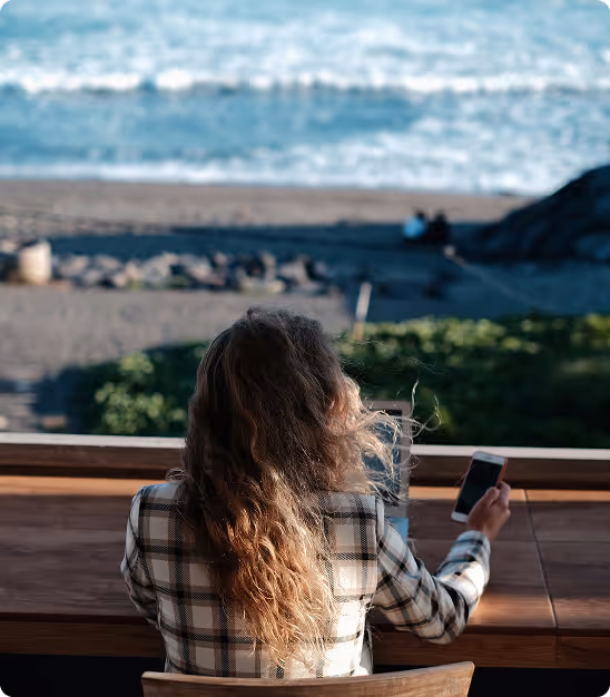 Femme assise devant une table en bois regardant la mer en tenant un smartphone.