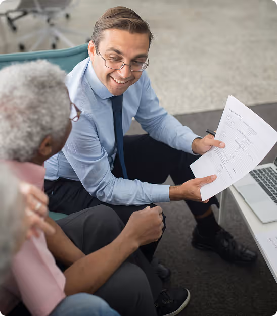 Un homme en chemise bleue et cravate souriant en montrant un document à deux personnes âgées en réunion.