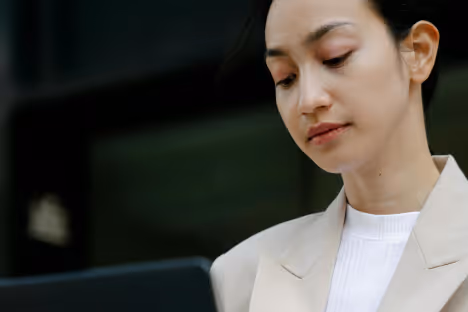 Une femme habillée d'un blazer beige regardant un ordinateur portable noir.