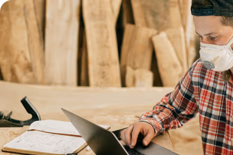 Person wearing a face mask and plaid shirt using a laptop in a woodworking workshop.