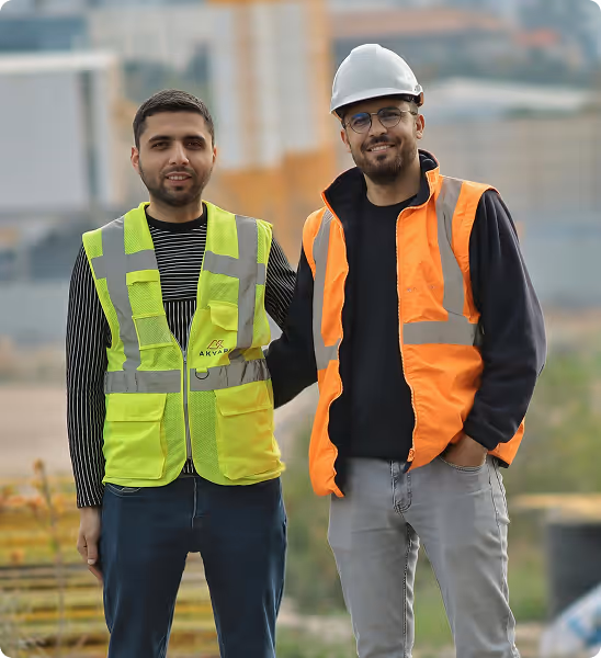 Deux hommes sur un chantier, l'un porte un gilet jaune fluo, l'autre un gilet orange avec un casque blanc.