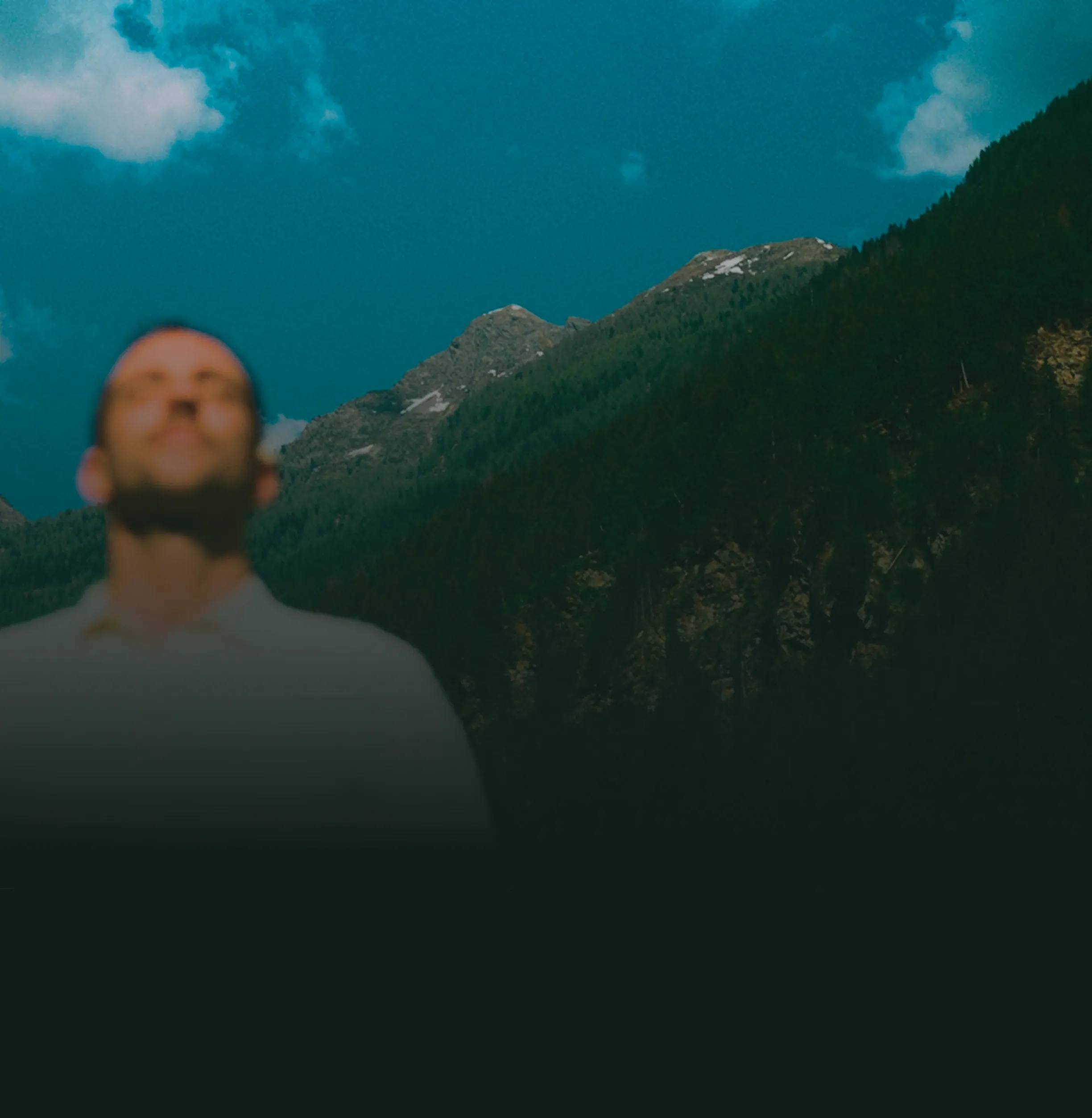 Homme flou les yeux fermés devant une montagne boisée sous un ciel bleu avec quelques nuages.