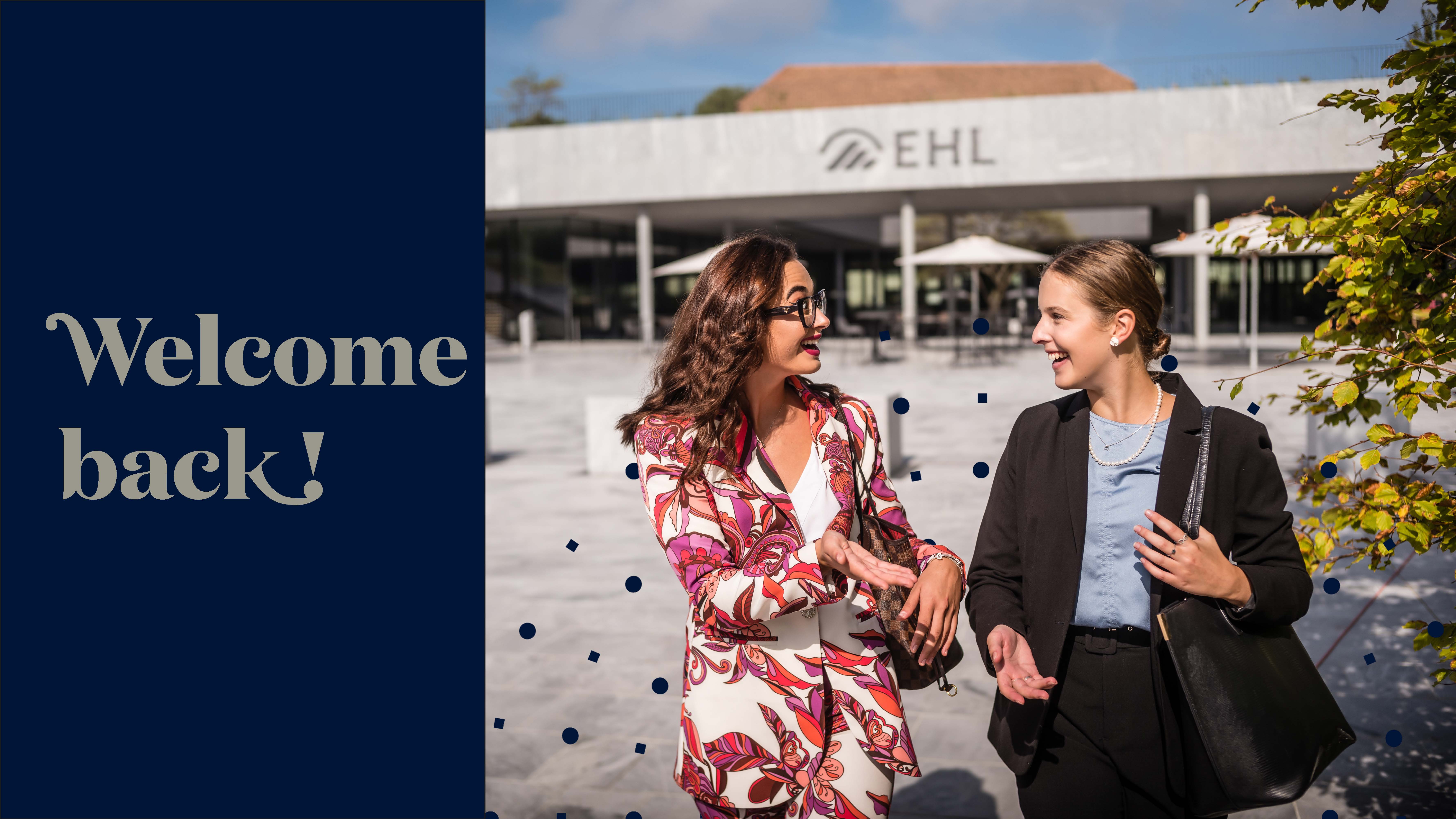 Deux femmes souriantes parlant devant un bâtiment avec le logo EHL sous un ciel bleu.