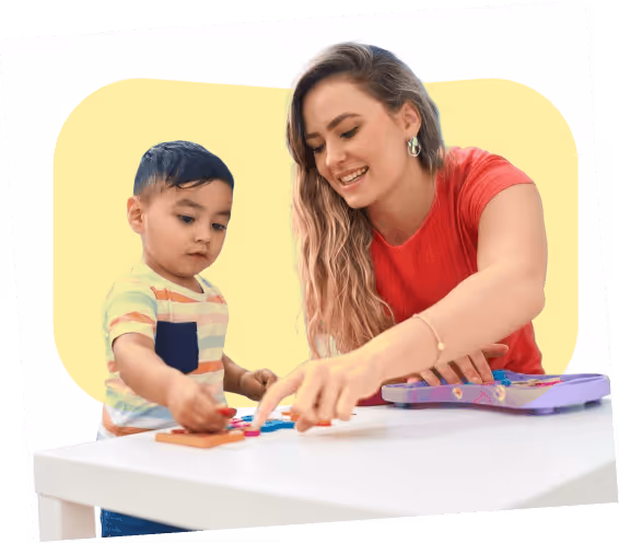 Young woman smiling while helping toddler boy with a puzzle at a white table.