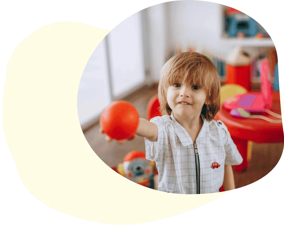 Young child with brown hair in a white checkered shirt holding and offering a red ball indoors.