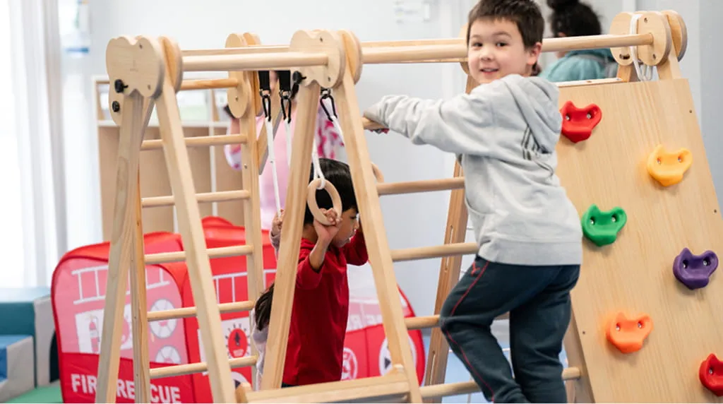 Two children playing on a wooden indoor climbing and swinging structure in a playroom.
