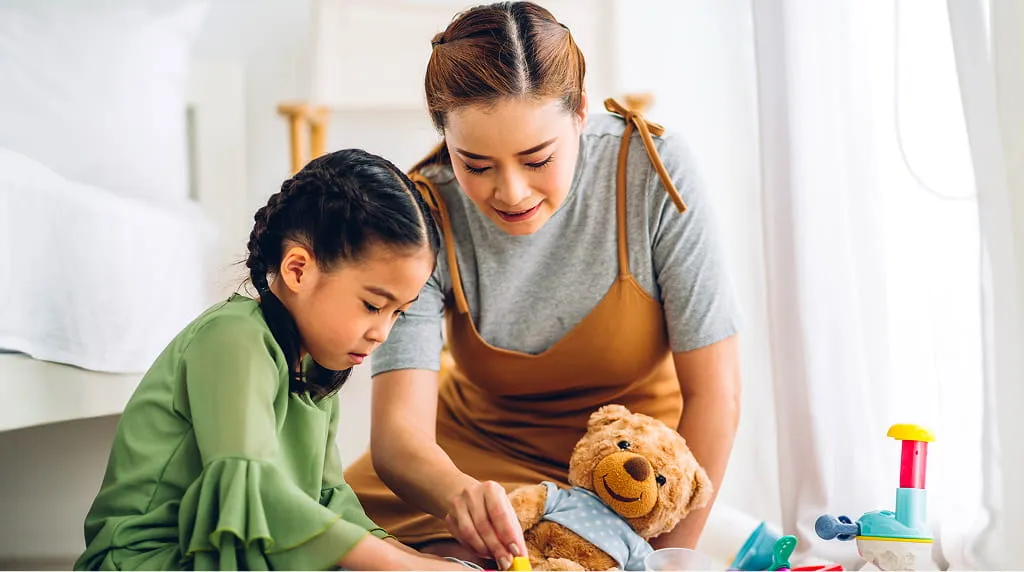 Woman and girl playing with toys and a teddy bear on the floor by a bed in a bright room.
