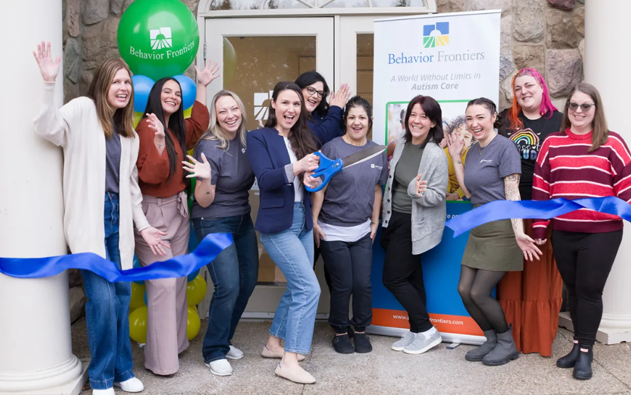 Group of nine diverse women cutting a blue ribbon celebrating Behavior Frontiers autism care center opening.