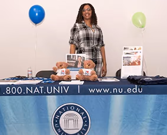 Woman in plaid dress standing behind a National University table with balloons, bears, and brochures.