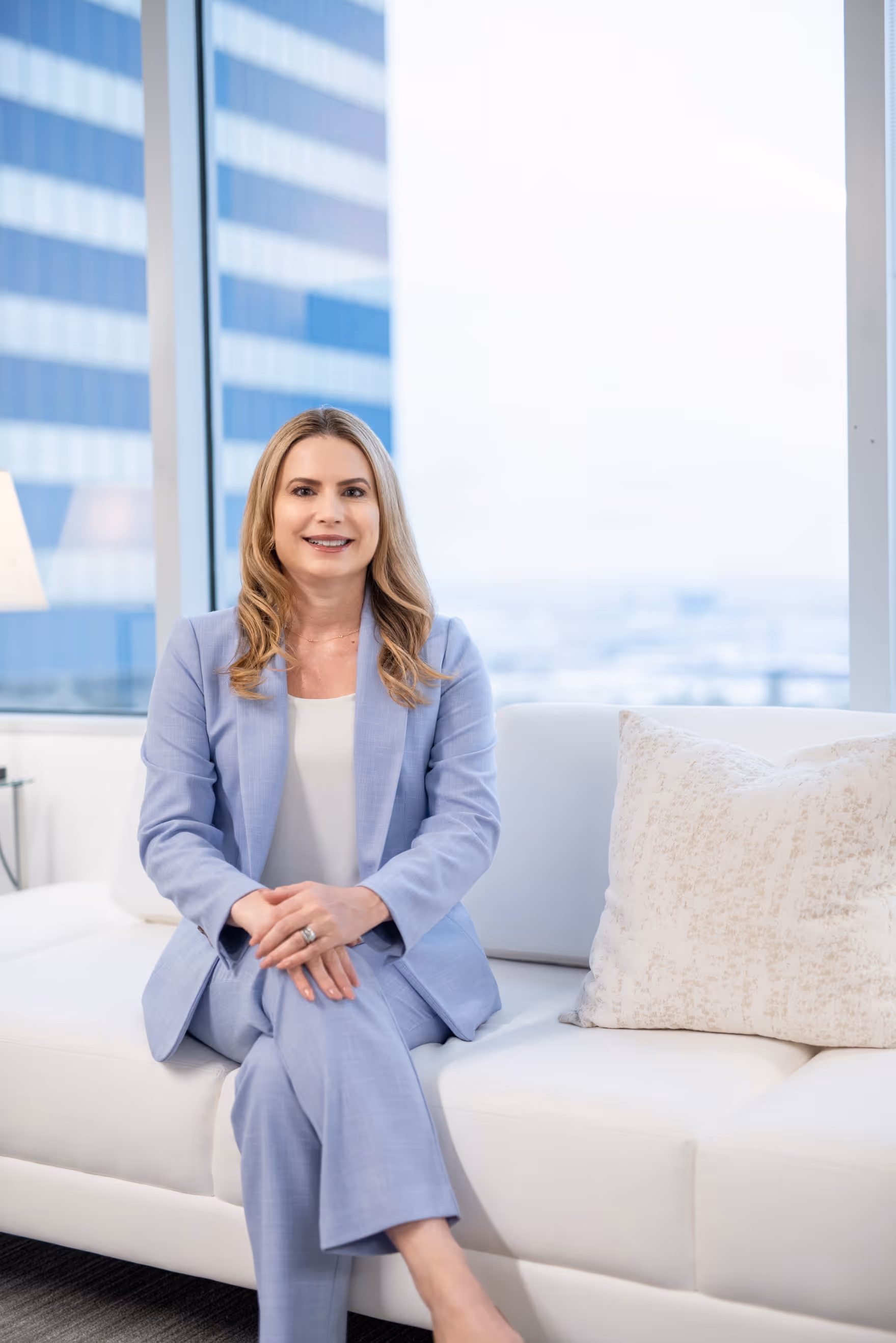 Smiling woman in a light blue suit sitting cross-legged on a white couch in an office.