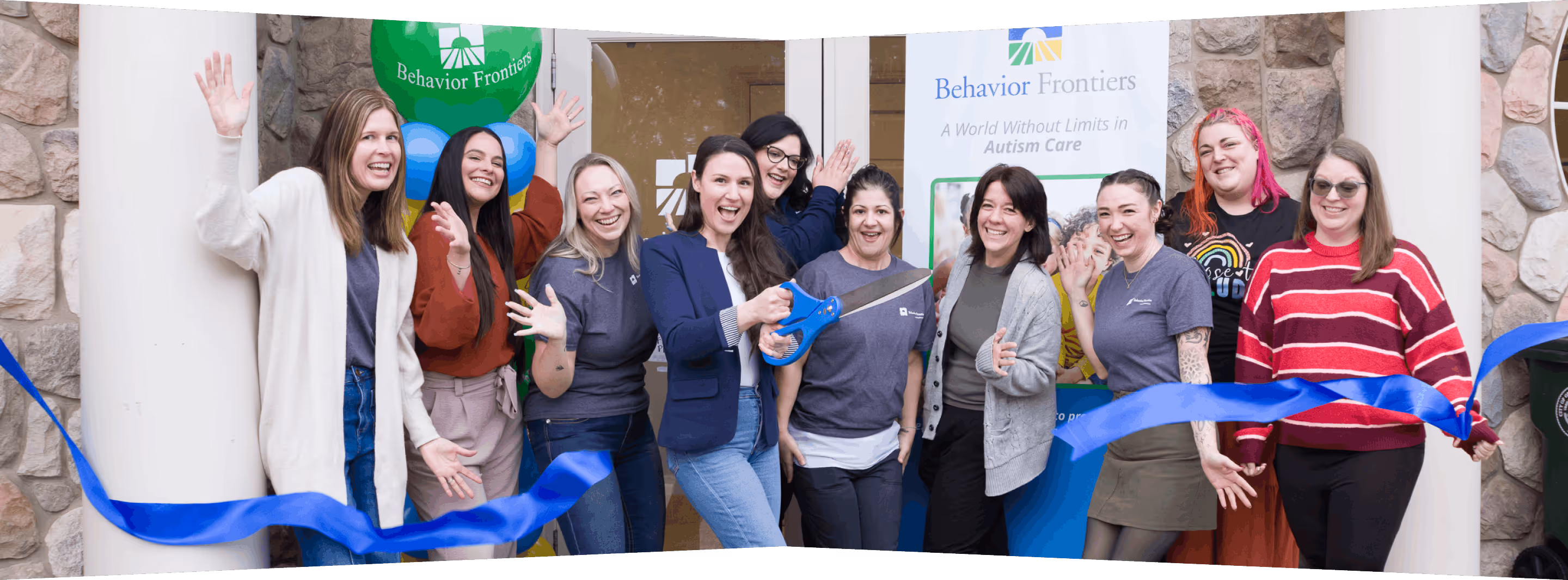 Group of people smiling and waving during a ribbon-cutting ceremony with blue ribbon and large scissors.