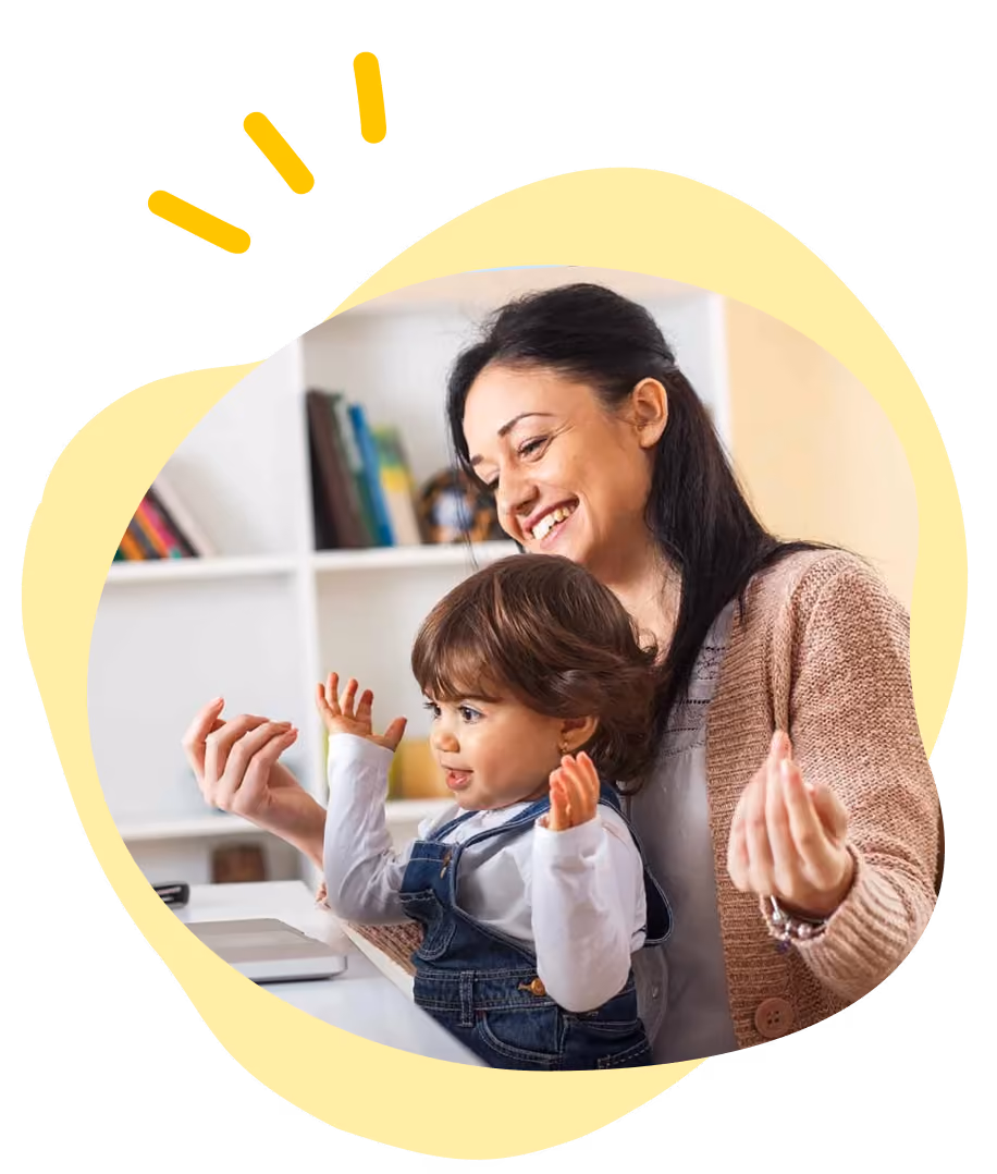 Smiling woman and child sitting at a table with raised hands in a bright room with shelves.