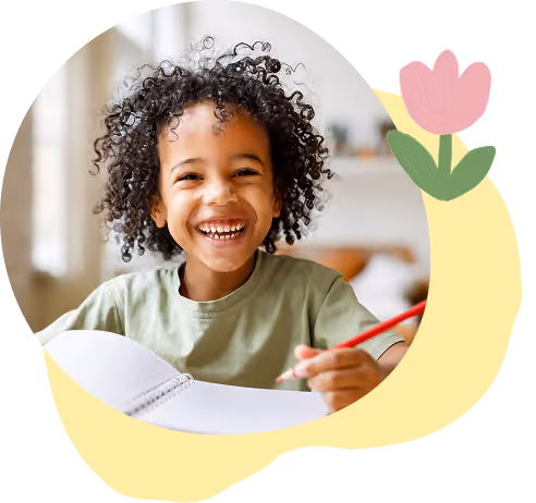 Smiling child with curly hair holding a red pencil and notebook at a table indoors.