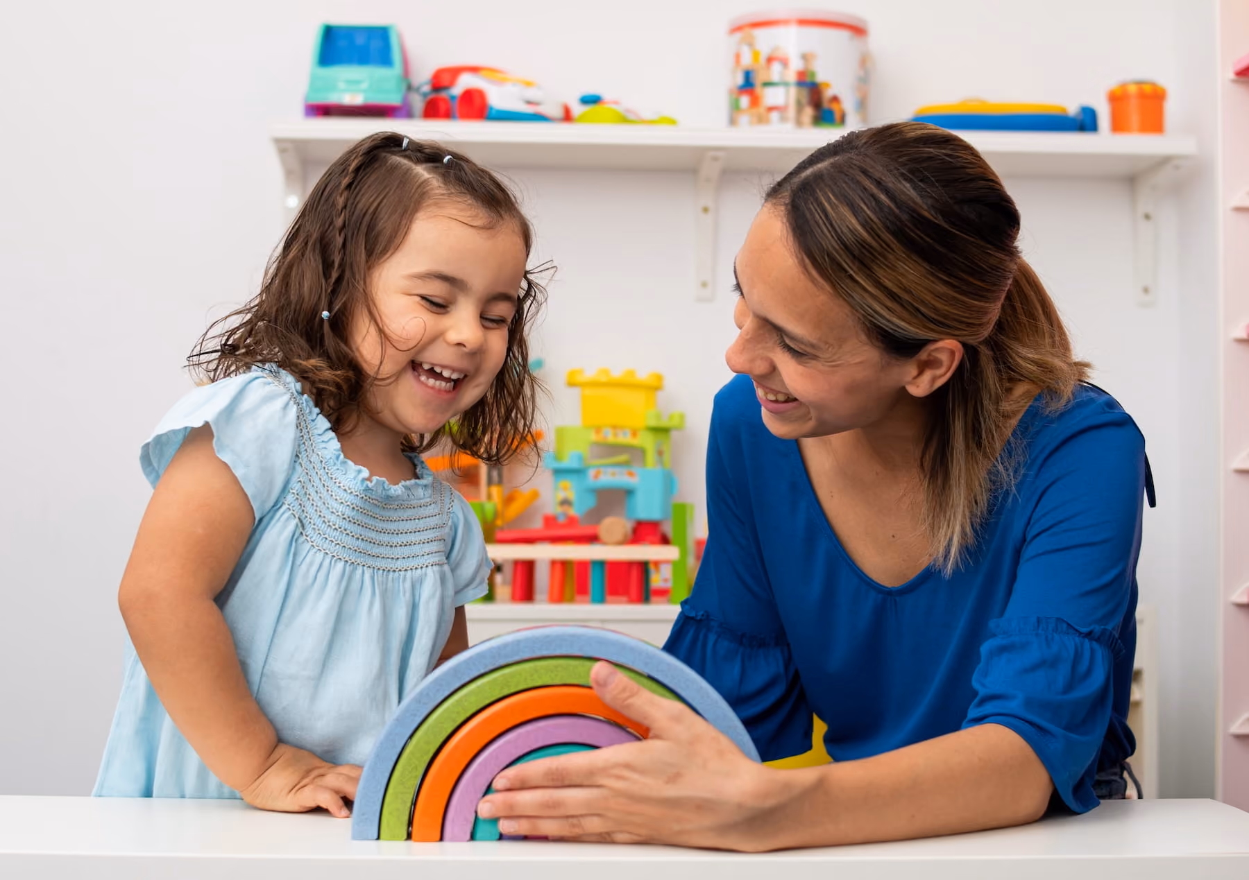 A laughing young girl in a light blue dress and a smiling woman in a blue top lean over a white table together, playing with a colorful stacked rainbow toy in a bright playroom with shelves of toys in the background.