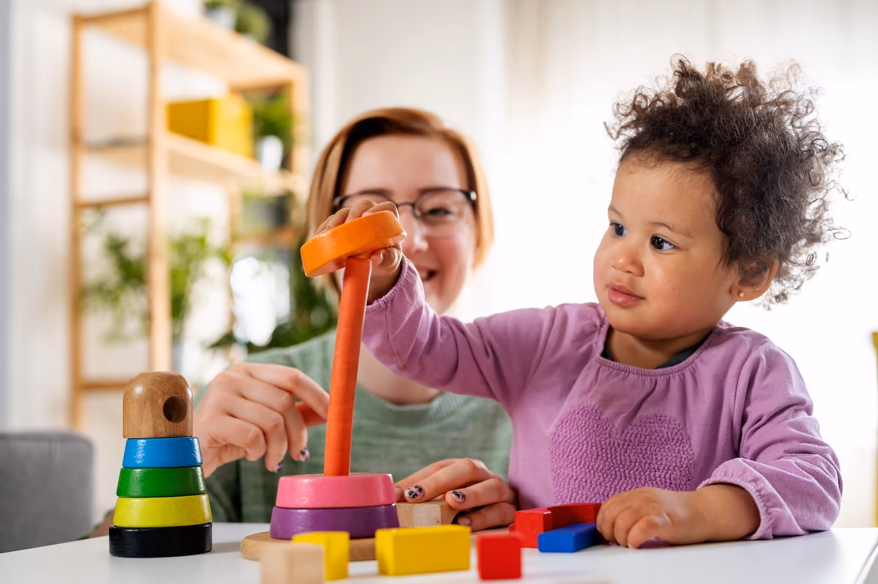 A smiling woman with glasses and red hair helps a toddler in a purple top stack colorful rings onto a wooden peg toy at a white table, with small wooden blocks scattered nearby and a bright, plant-filled room in the background.