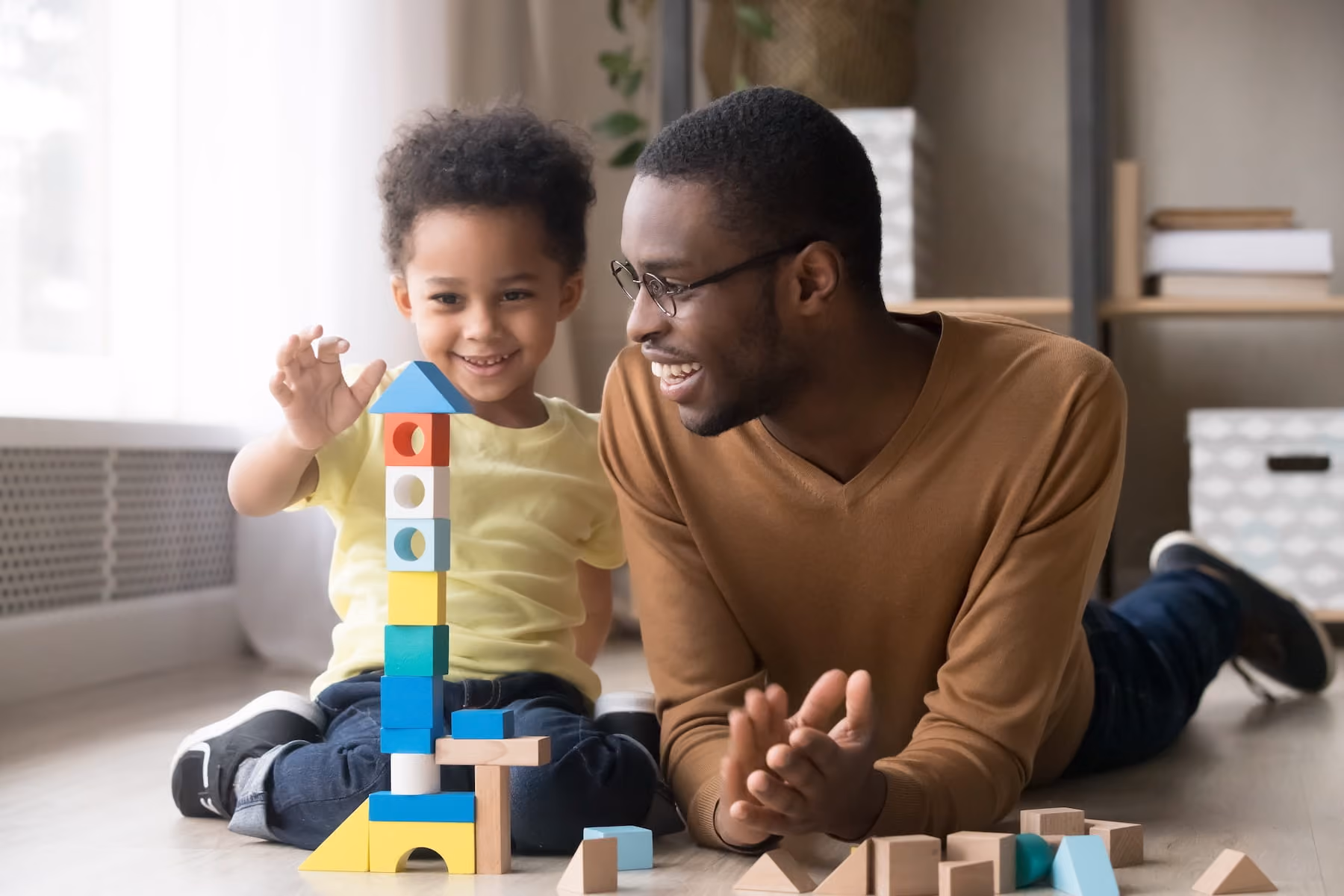 A smiling young boy in a yellow shirt proudly shows off a tall tower he built from colorful wooden blocks while a laughing man in a brown sweater and glasses lies on the floor beside him, cheering him on in a warm, cozy living room.