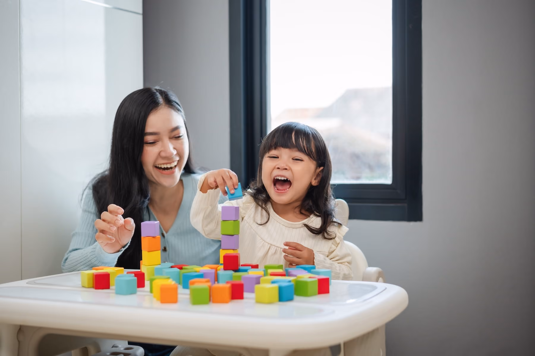 A laughing young girl and a smiling woman sit together at a small white table covered in colorful square blocks, joyfully stacking them into towers near a bright window.