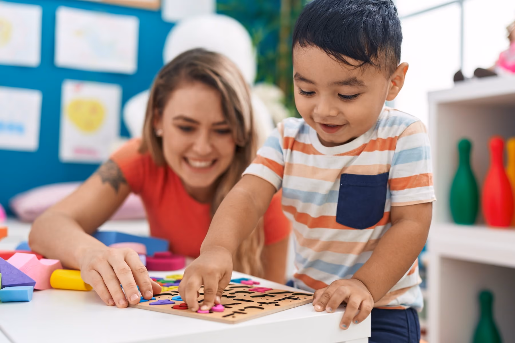 A smiling woman in a red top and a young boy in a striped shirt work together on a colorful wooden letter puzzle at a table in a bright, toy-filled classroom.