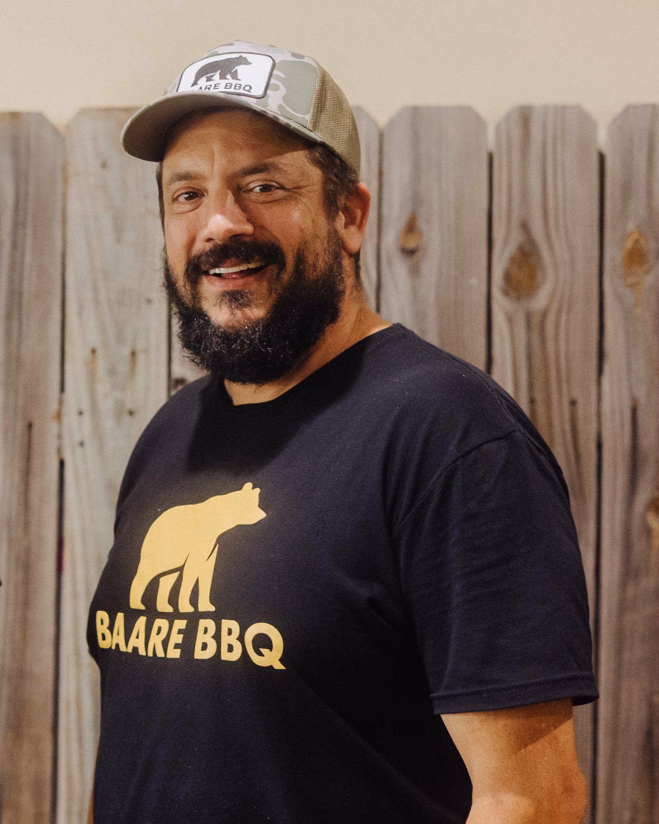 Baare BBQ owner Bret wearing camouflage cap and a black t-shirt with a yellow bear and the text 'BAARE BBQ' in front of a wooden fence.